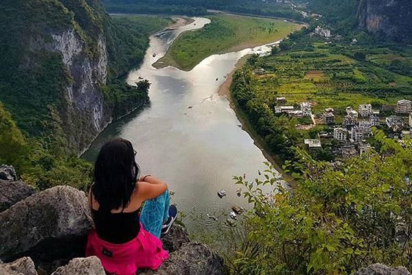 A study abroad student sits on a rock, facing over a valley with a river snaking through it.