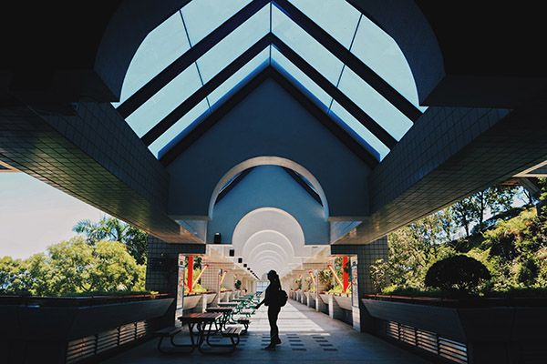 A person stands on a darkened bridge with a roof, and light pouring in from outside creating shadows. There is a row of benches next to them.