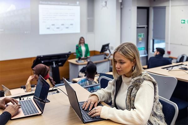 Students in a lecture theatre using laptops while a lecturer presents at the front with a projected slide.