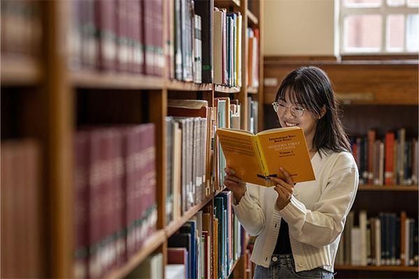 Student standing between library bookshelves, smiling while reading an orange book.