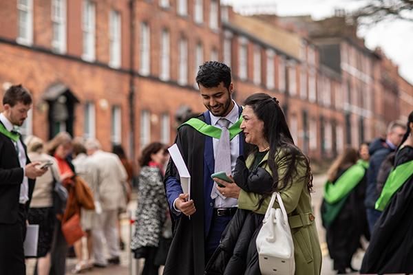 A graduate in a gown with their guest on graduation day in front of a red brick building om campus