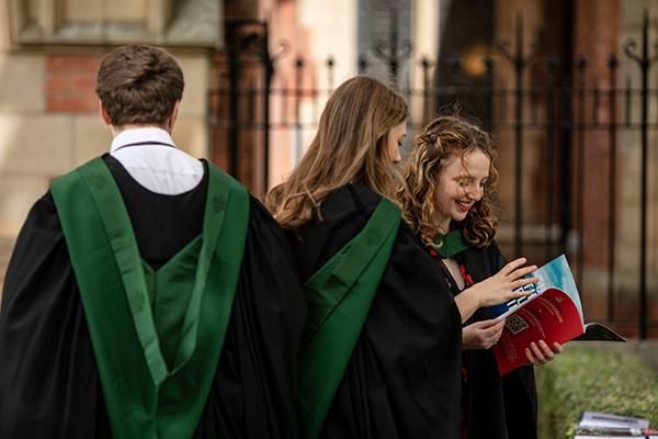 Three graduates in gowns, one is reading their graduation programme