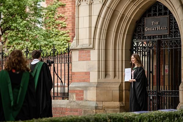 Graduate in a gown holding their dissertation for a photo outside the Great Hall