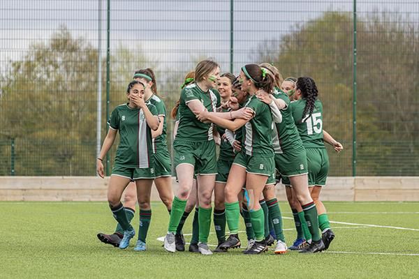 A group of Gryphons football players wearing green kits gather closely together celebrating on an outdoor pitch
