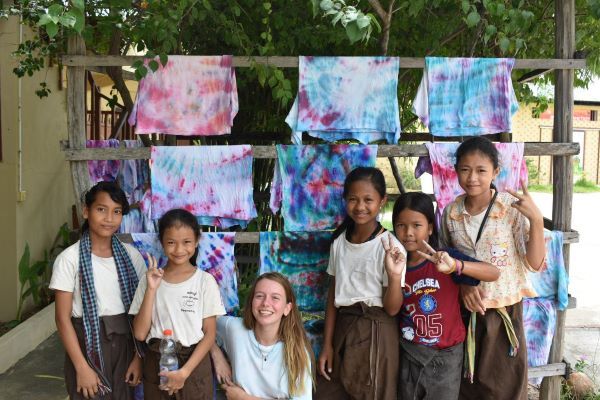 A student crouches smiling with a group of children. Behind them are tie-dyed pieces of fabric, hanging up.