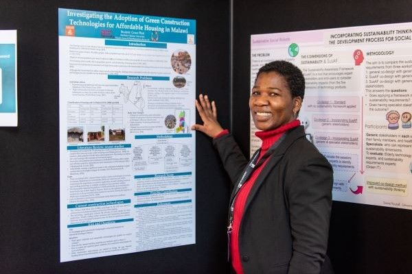 A student stands facing the camera, pointing towards a research poster on the wall.
