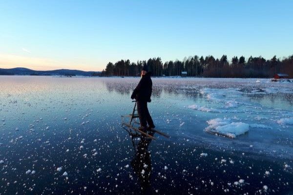 A student stands on a sheet of ice, dappled with snow. They are holding a wooden sled and behind them is a clear blue sky and a landscape of trees.