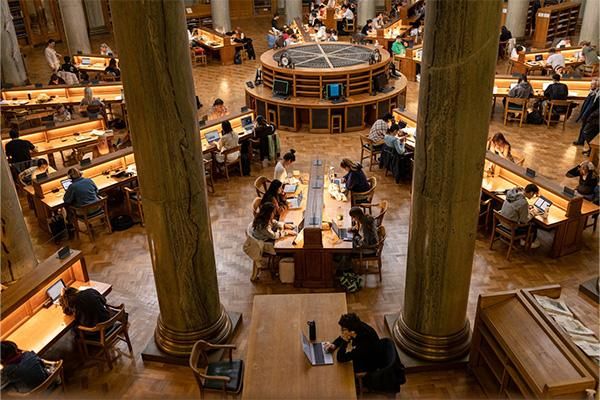 Large library reading room with tall columns and rows of wooden desks, where many students are studying with books and laptops.