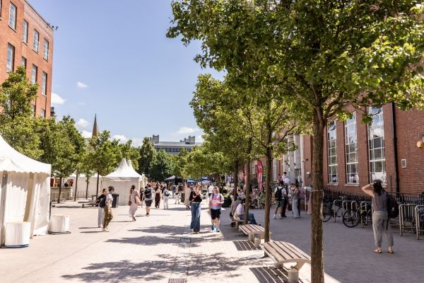 Rows of white event tents line University Precinct, while numerous people are walking, browsing stalls, meeting others, or sitting at nearby café tables.