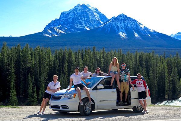 A group of students stand in and around a mini van, parked in front of a huge mountain with trees below.