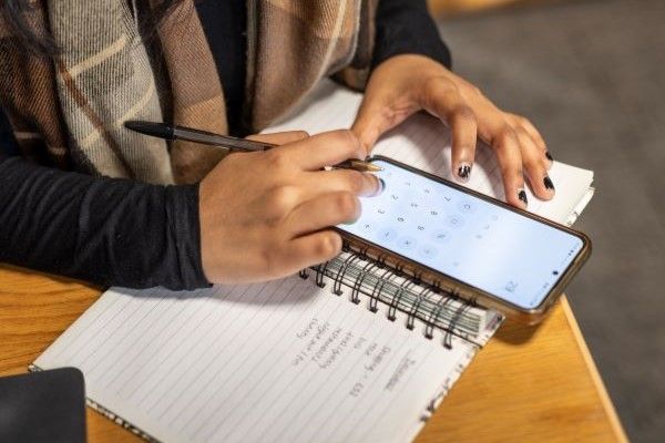 A student using a calculator app on their phone while holding a pen and taking notes on paper.