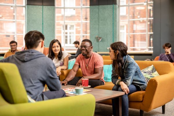Four students chat in a café at LUU. Three face the camera, looking toward the fourth, who sits opposite. They're on orange and green sofas, with hot drinks on the table and large windows in the background.
