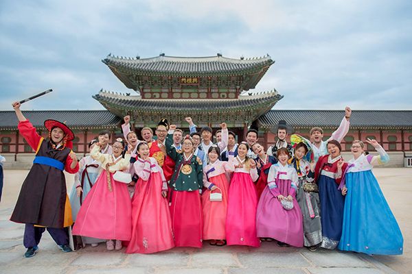 A group of people stand in traditional Korean dress, smiling at the camera with arms outstretched. Behind them is a traditional Korean building.