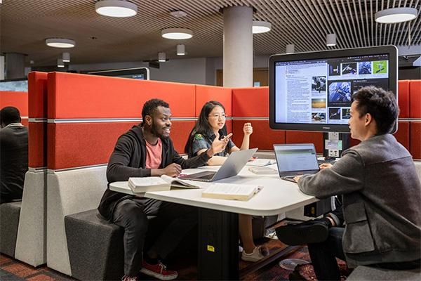 Three students sitting in a modern study space with laptops and books, working together around a large screen display.
