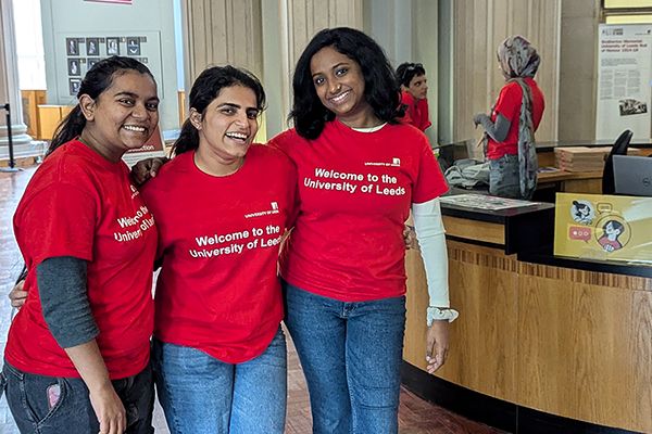 Three smiling student ambassadors wearing matching red "Welcome to the University of Leeds" t-shirts stand together at the Welcome Hub