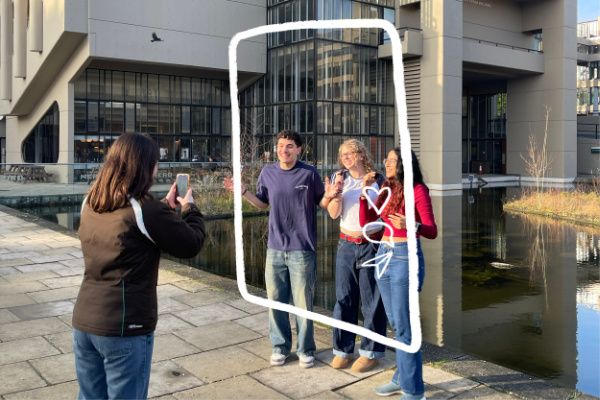 A student filming a trio of students next to the pond outside Roger Stevens building. The trio are framed by a hand-drawn phone screen graphic.