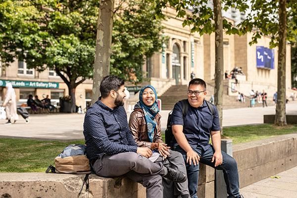 Three students sit together on a stone bench outdoors, laughing and talking. In the background is the large, classical-style facade of the Leeds Art Gallery on a sunny day.