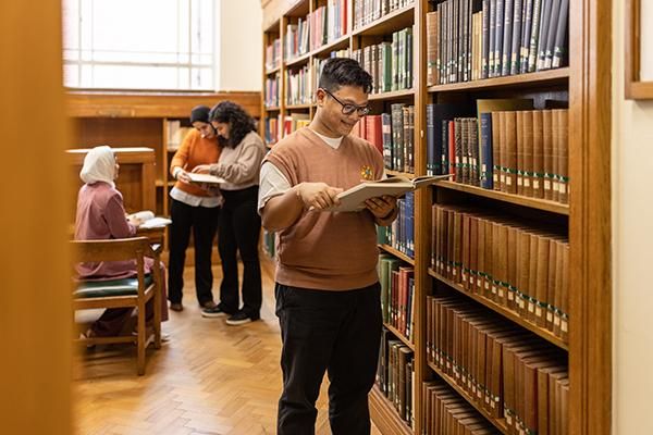 Student reading book in a library next to shelves of books