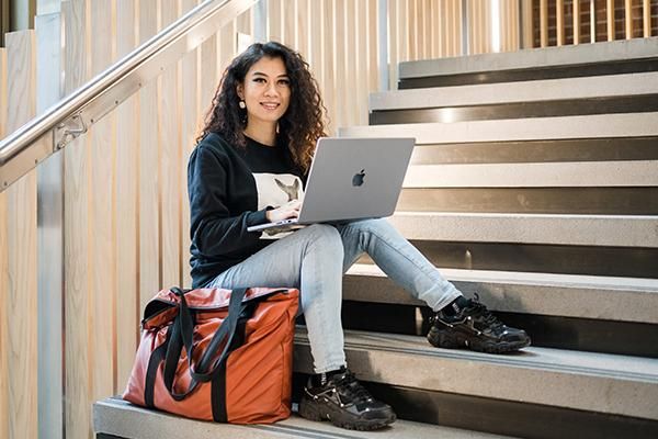 A student sitting on some steps with a laptop on their knees and a large orange bag next to them