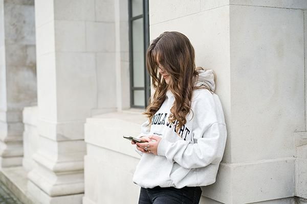 Student wearing a hoodie leaning against a wall using their phone