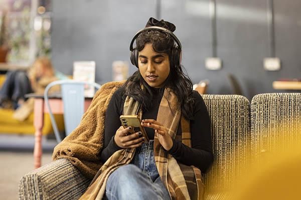 Student sitting on a sofa wearing headphones and scrolling on their phone