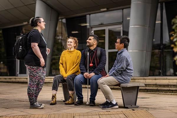 Three students sitting on a bench chatting to another who is standing