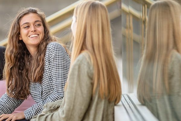 Two friends sat on steps on campus chatting. One is smiling at the other.