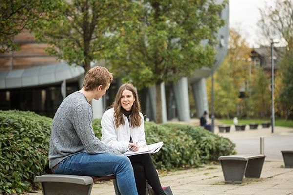 Two students talking on a bench outside the Business School. One has a notepad.