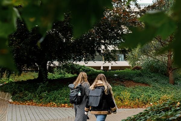 Two students walk along a paved area surrounded by bushes, shrubs and trees