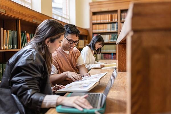 Three students sitting at a wooden desk in a library with shelves of books behind them. Two students in the foreground are smiling and looking at an open book together, while a laptop sits nearby. A third student in the background is focused on reading.