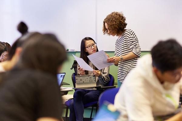 A tutor holding a piece of paper is supporting a postgraduate student in a class