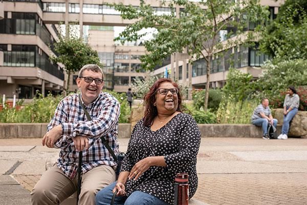 Two mature students sitting on campus laughing surrounded by buildings and trees.