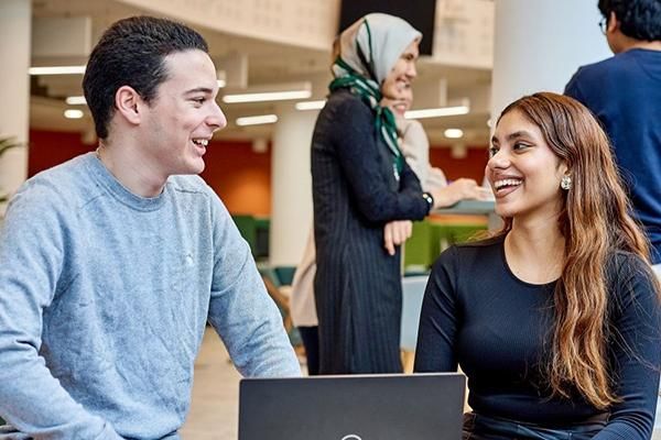 Two people sit and chat. A laptop is in front of them on a table