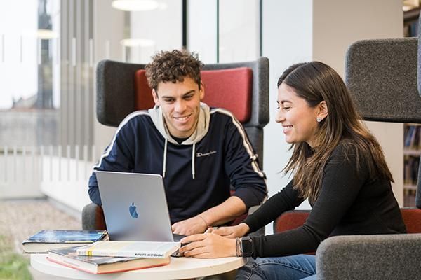 Two students sit in modern armchairs in a library, smiling as they look at a laptop screen together. Books are on the table in front of them, suggesting collaborative study.