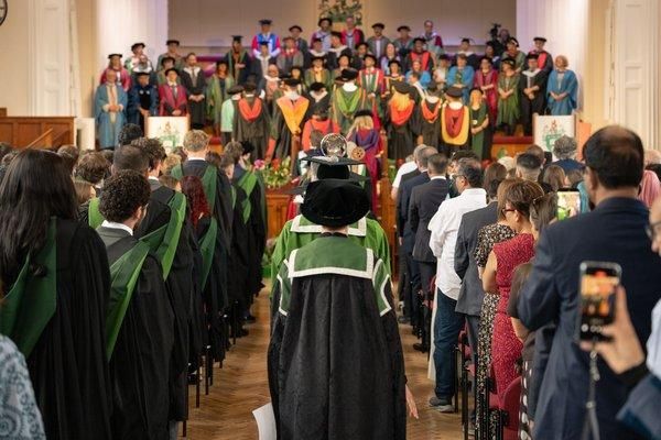 Graduates and guests stand as the academic procession, led by the mace bearer, walks down the aisle towards the platform party.