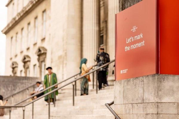 A red Grad 2026 sign reading “Let’s mark the moment” beside the Parkinson Building steps, with graduates and guests in the background.