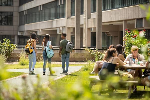 Students walking across campus with backpacks while others sit at picnic tables in a grassy outdoor area.
