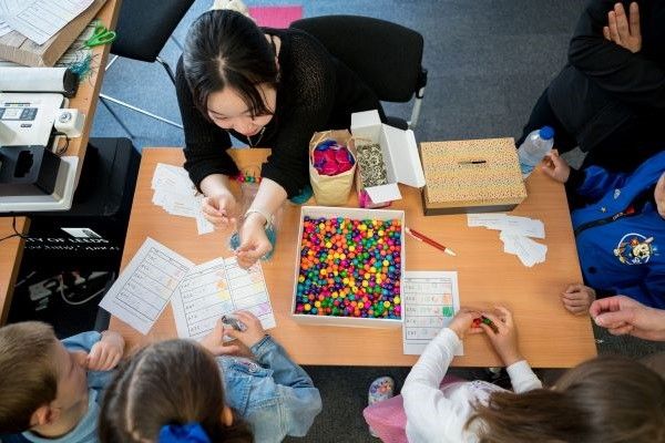 Close up shot, taken from above, of an adult's and children's heads and hands as the adult shows the children what they need to do for an activity at a stall. There's a box of different coloured beads, which can be threaded onto small metal hoops and stri