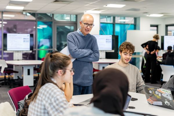 An academic tutor stood teaching in the middle of a large, bright classroom, talking to a table of students who are working on their laptops.