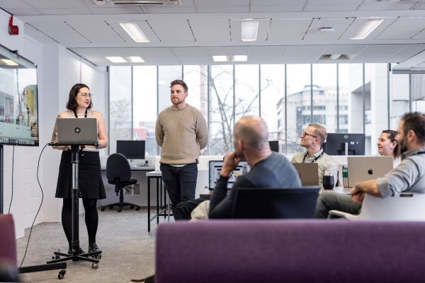 Two people stand and present at a meeting, whilst other people sit around a table and listen. One of the presenters is standing behind a portable lectern and there is a large screen beside them.