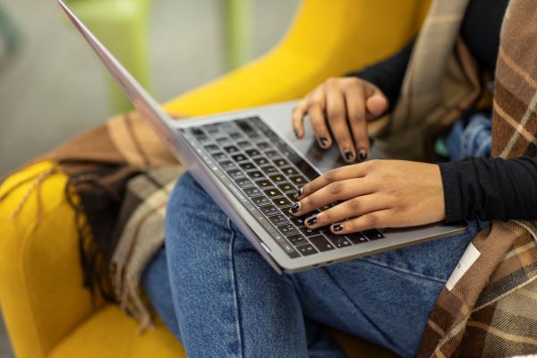 Close-up of a student’s hands as they sit comfortably in a yellow, upholstered chair and type on a laptop.