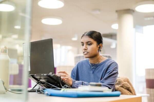A student, who's wearing a blue sweater and bluetooth earphones, sits and works on their laptop inside a library on campus during the day.