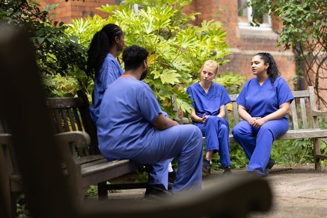 A small group of people, wearing blue medical scrubs, sit outside on wooden benches inside a courtyard and chat casually. The courtyard is surrounded by red brick buildings and there are green shrubs in the background.