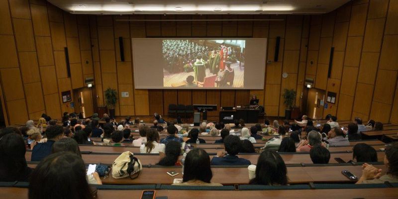 Guests seated in a lecture theatre watch a live stream of the graduation ceremony on a large screen.
