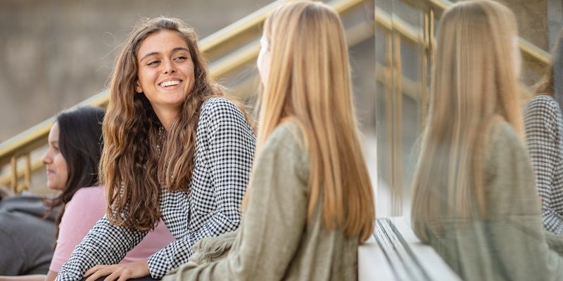 Two friends sat on steps on campus chatting. One is smiling at the other