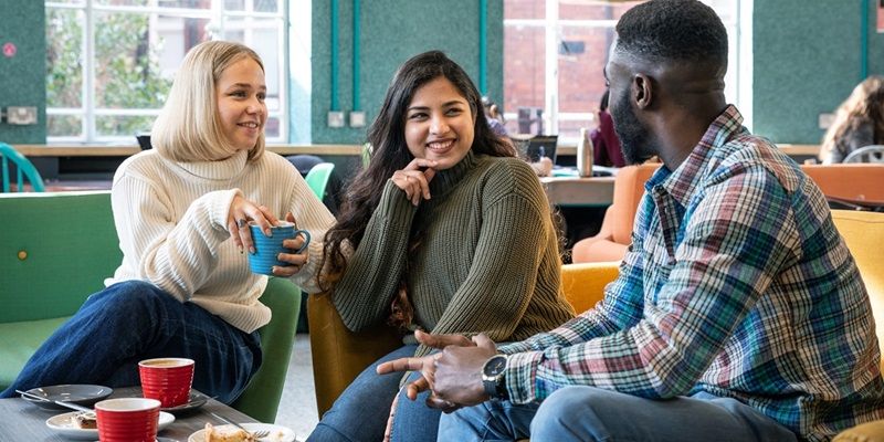 Three students sat in a bright comfy social space, drinking coffee and chatting