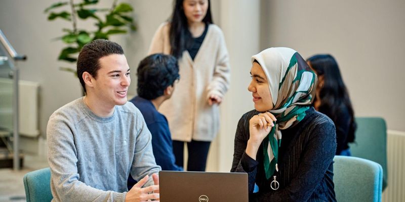 Students sit at a table and chat whilst working on a laptop.