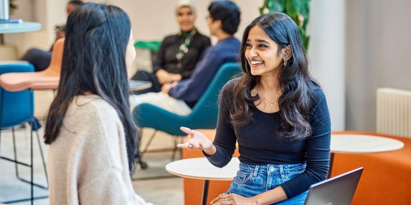Students sit at a table and chat whilst working on a MacBook