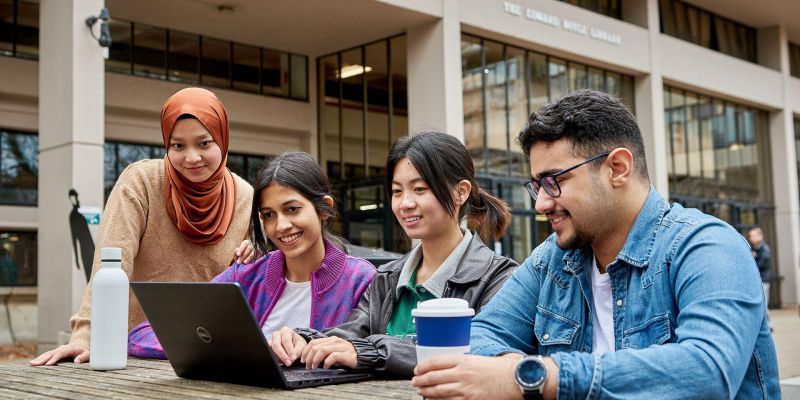 A group of students working together on a laptop on a wooden table outside the Edit Room café on campus.