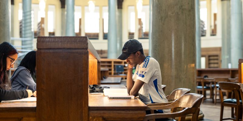 Students sit at long wooden tables in a spacious academic library, reading and studying independently.